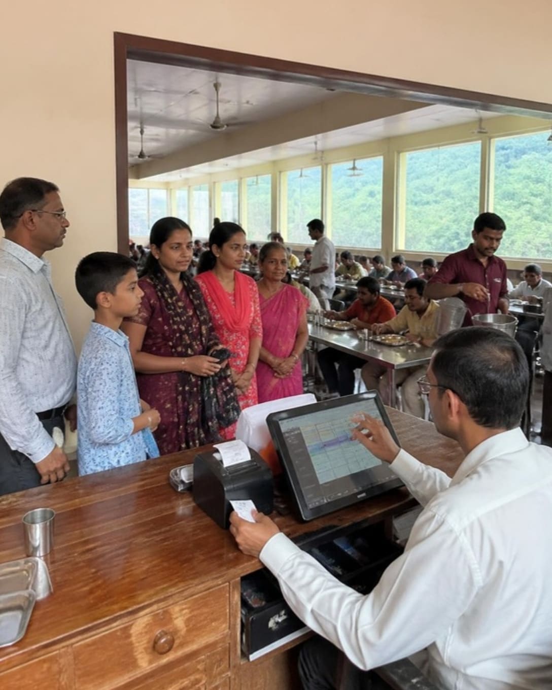 A cashier at the entrance of a dining hall billing thalis on a touchscreen POS and handing a token to a pilgrim family